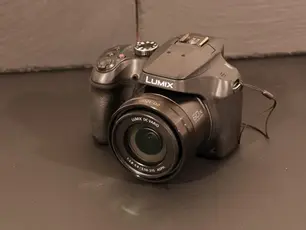 A black Lumix camera with "60X" and "LUMIX DC VARIO" text on the lens rests on a dark surface, angled against a neutral background.