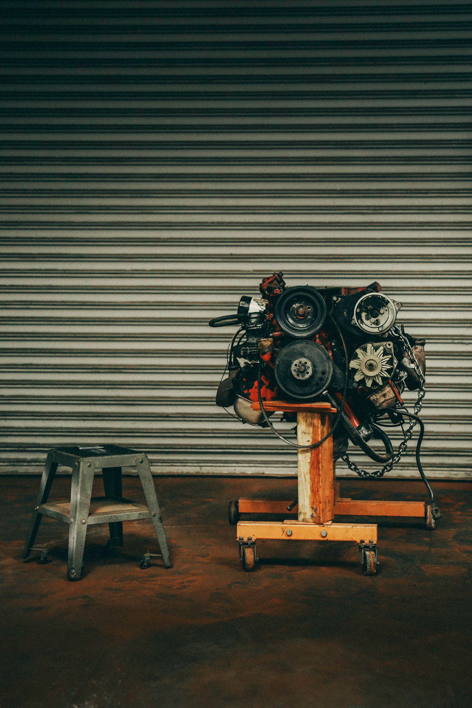 Classic car engine with pulleys and belts mounted on wooden engine stand in Los Angeles restoration garage with corrugated metal wall and step stool