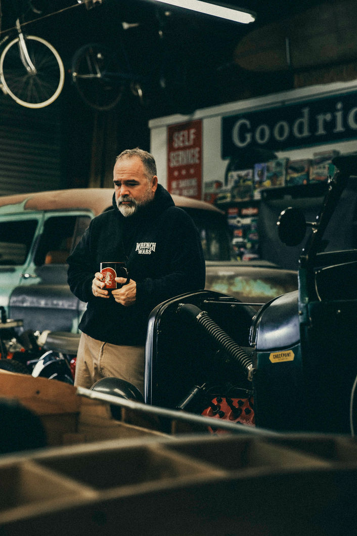 Custom builder Mark Jarel holding coffee mug in workshop with vintage motorcycles and cars in Torrance California garage
