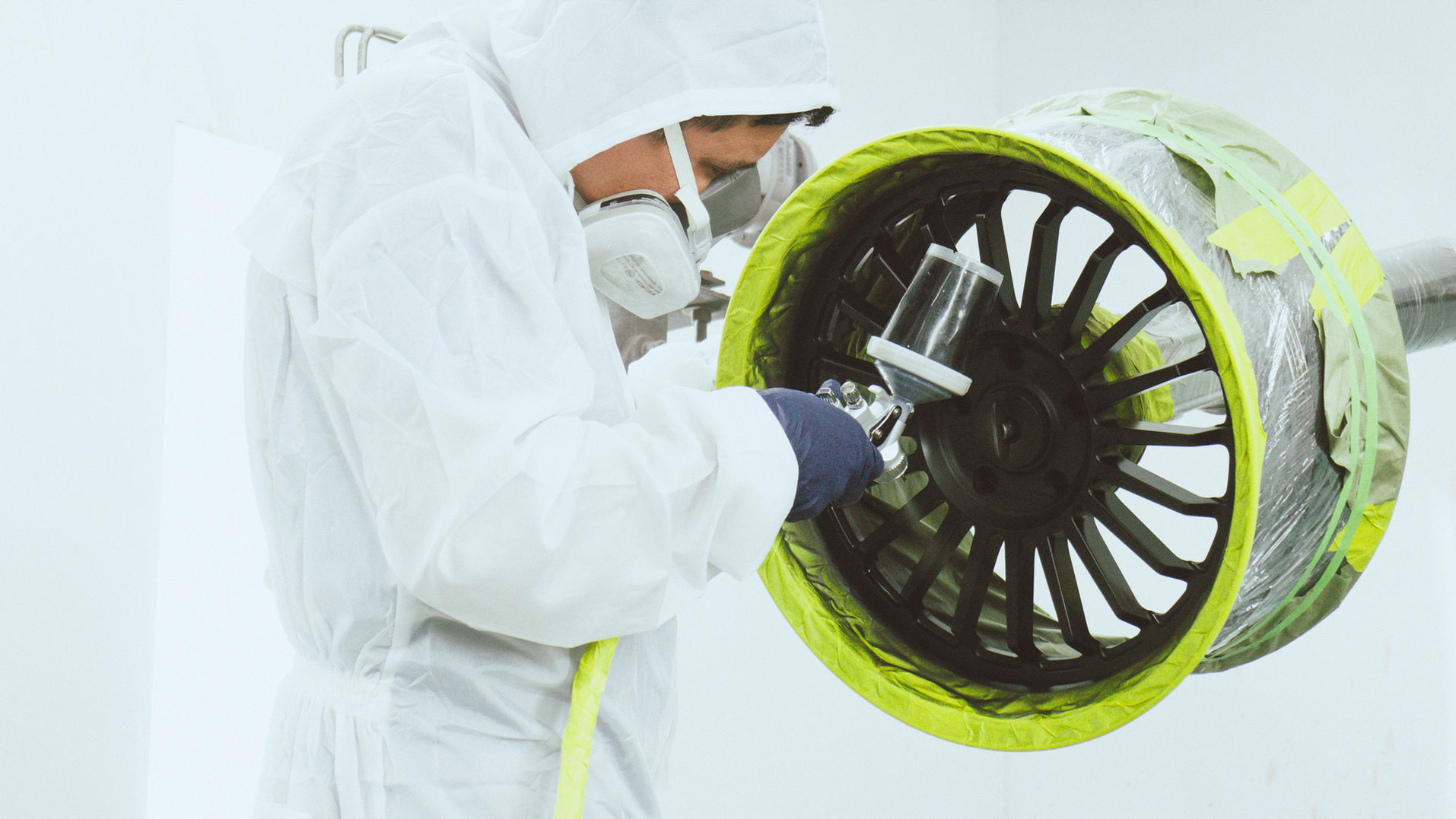 Breithaus Wheels crew member in protective suit spray painting black wheel with yellow masking in professional paint booth facility