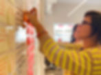 Two middle-aged women place sticky notes on a white wall covered with yellow and pink notes in a shiny room with a big window in the background. The one at the front, tanned with brown, wavy, medium-length hair and glasses, is wearing a striped yellow and brown sweater and a beaded multicolour bracelet. The one at the back is white with curly black hair and a white shirt.