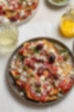 Greek dakos salad on a plate with feta, olives, and capers. Olive oil and a glass of white wine are beside it on a white tablecloth.