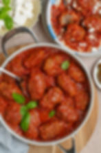 Meatballs in rich red tomato sauce garnished with basil in a pan; feta on a plate nearby. Rustic wooden surface setting.