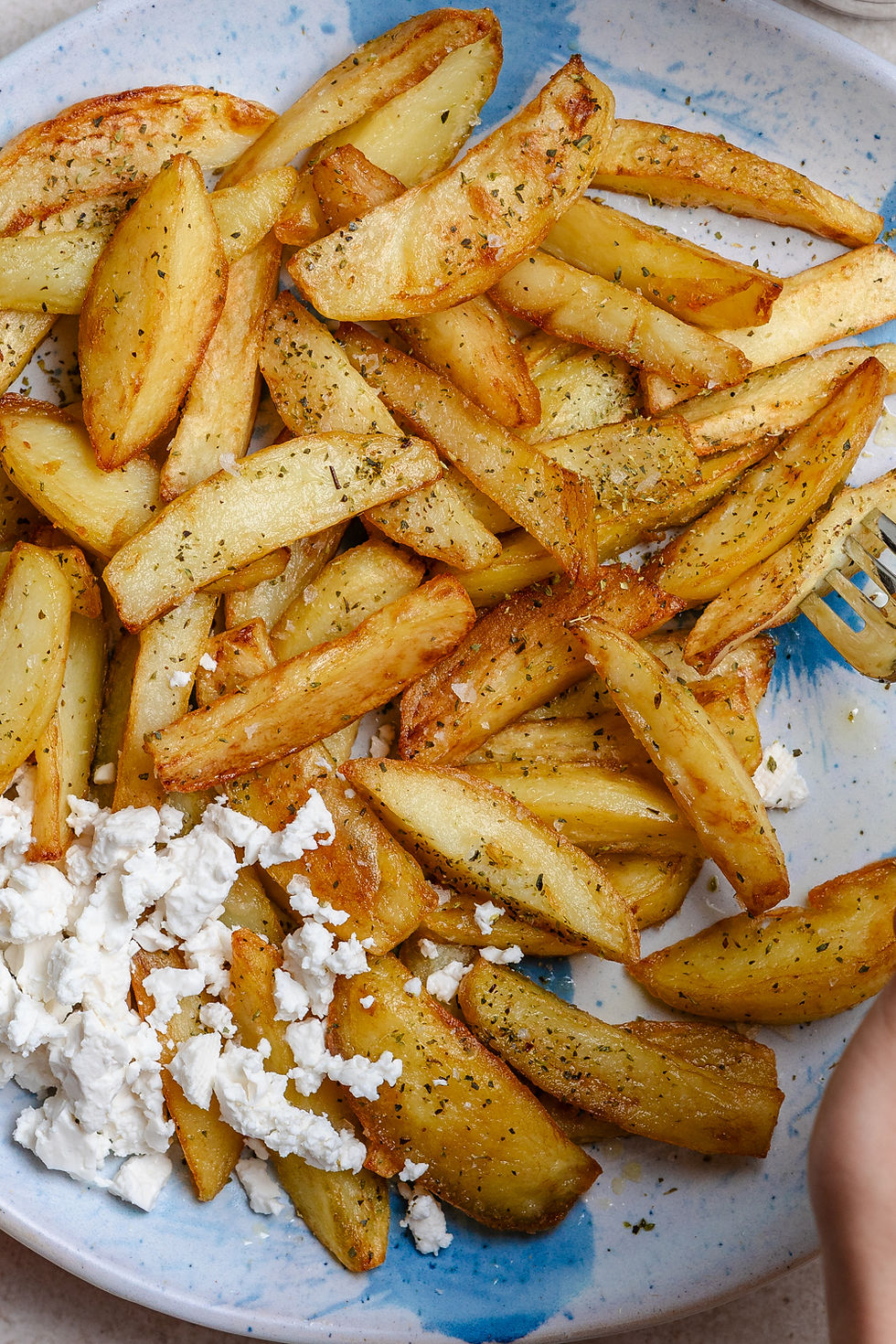 Roasted Greek Fried Potatoes (Patates Tiganites) with herbs and crumbled white cheese on a blue-speckled plate, with a fork at the edge.