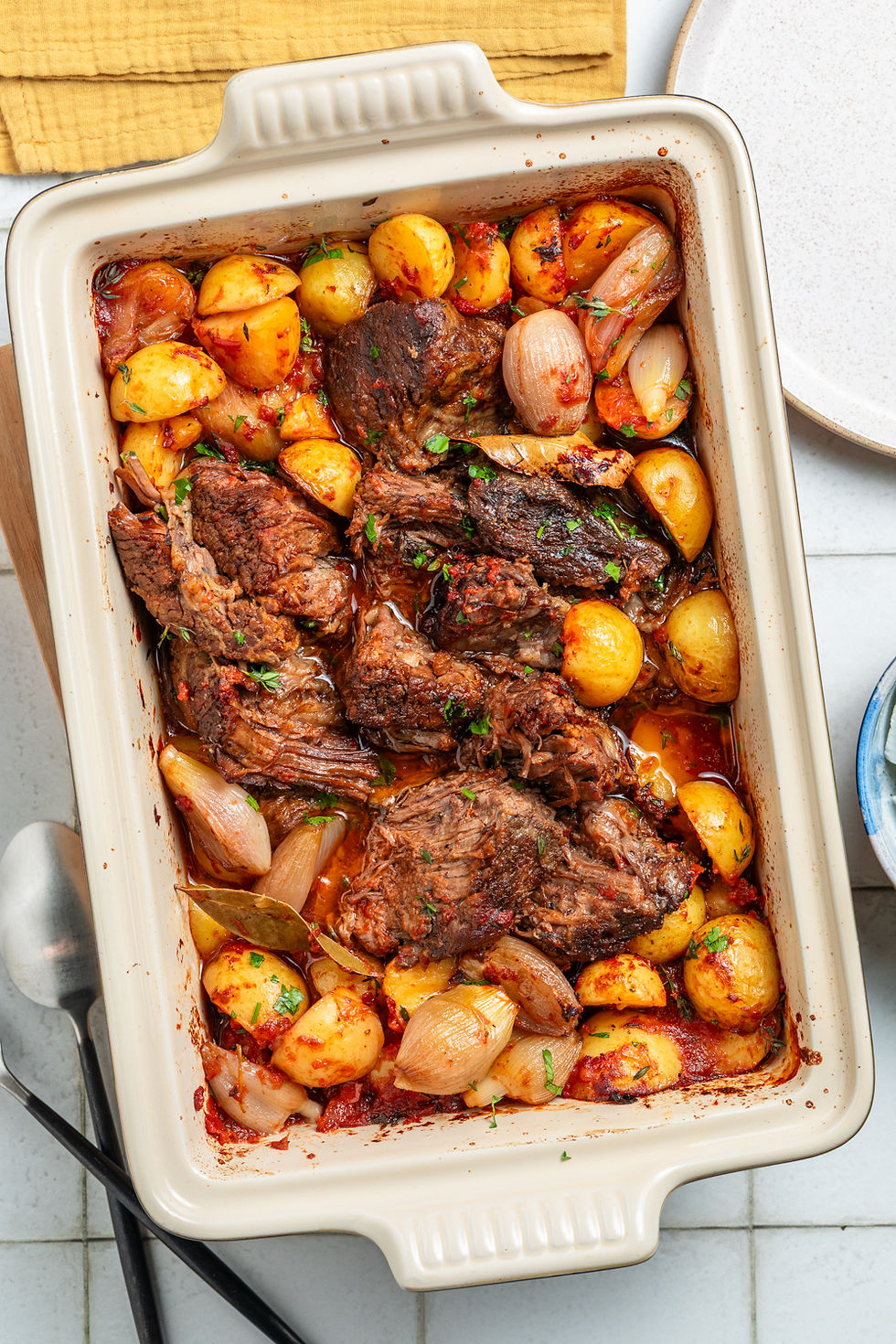 Roasted beef and potatoes in a rectangular dish, garnished with herbs, surrounded by onions. Set on a white tile surface with cutlery.