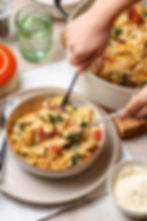 Hands serve creamy farfalle pasta with chicken, spinach, and sun-dried tomatoes into bowls on a cozy table.