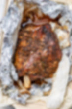 Roasted lamb with herbs on parchment in a tray, wrapped in foil. Juices visible, creating a rustic and appetizing presentation.