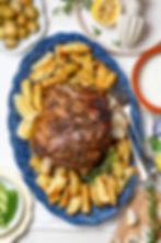 Roasted lamb with potato wedges on a blue platter, garnished with lemon and rosemary. Surrounding are olives, sauce, and cucumbers.