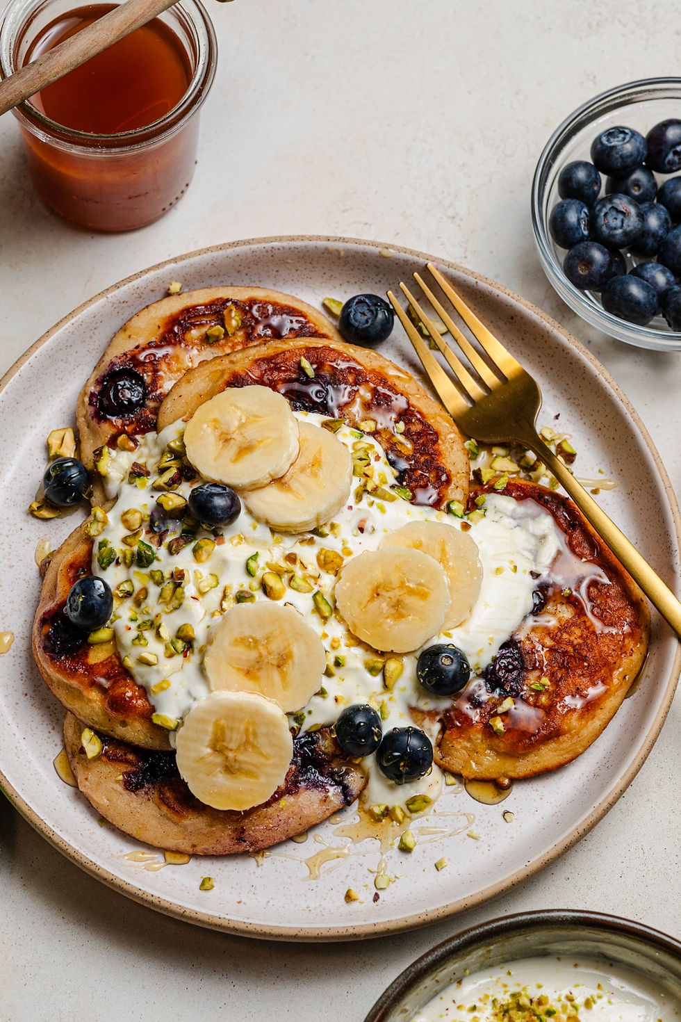 Pancakes topped with banana slices, blueberries, yogurt, and pistachios on a plate with a golden fork. Honey jar and blueberries nearby.