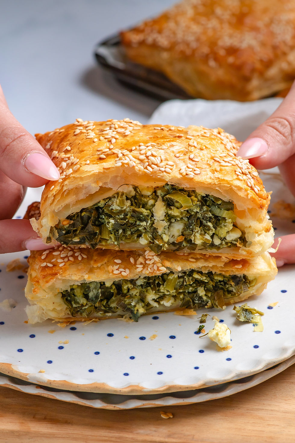 Spanakopita Pockets - Greek Spinach & Feta Pastries. Hands hold a sesame-topped flaky pastry cut open to reveal a green herb and cheese filling on a white polka-dot plate.