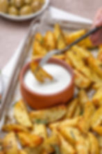 Hand dips roasted potato wedges in creamy white sauce on a tray. Olives in the background. Earthy tones, inviting scene.