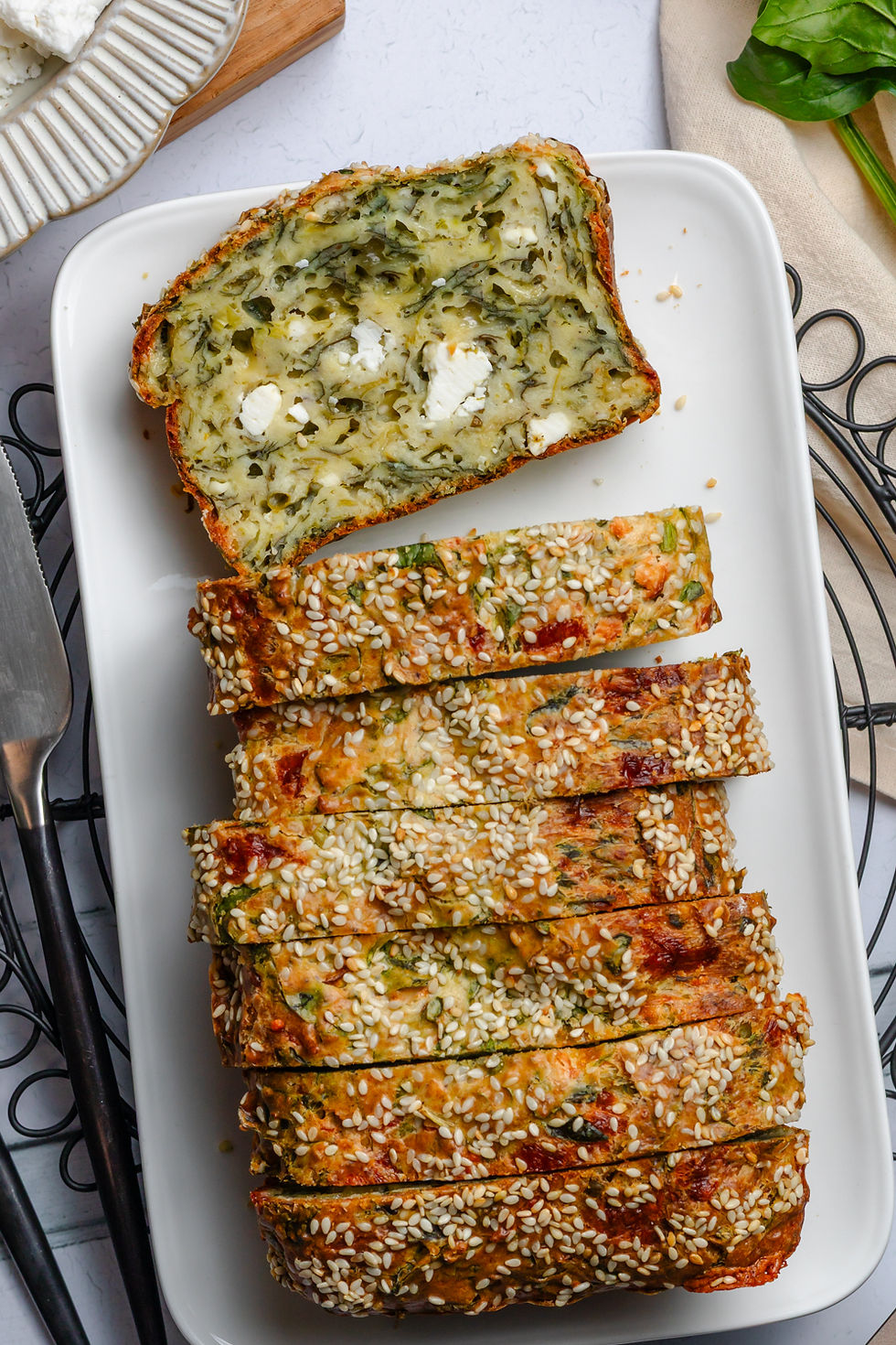 Sliced sesame-topped savory spinach and feta loaf and herb bread on a white platter, served overhead with rustic kitchen props and napkins
