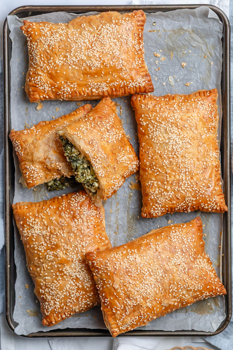 Spanakopita Pockets - Greek Spinach & Feta Pastries. Tray of golden sesame-topped pastries, one cut open to reveal spinach filling, on parchment-lined baking sheet