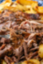 Close-up of tender shredded roast lamb with crispy crust on a fork, surrounded by roasted potatoes. Warm, rustic tones dominate the image.