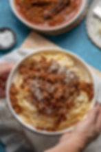 Hands hold a bowl of spaghetti topped with meat sauce and grated cheese. A dish of sauce and salt are in the blue-toned background.