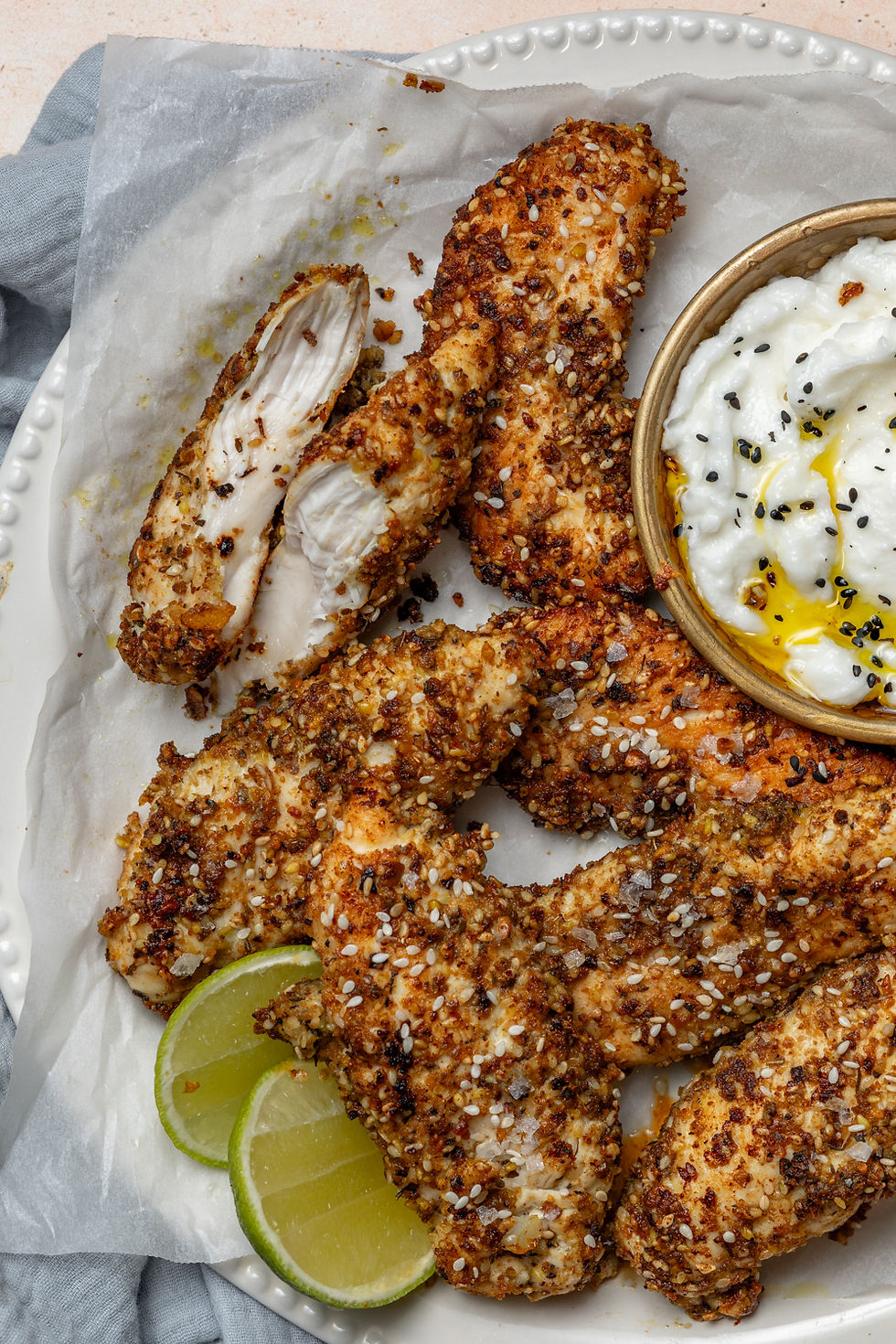 Crispy dukkah chicken tenderloins on a plate with sliced lime and a bowl of garlic dip. Set on parchment, evoking a zesty, savory mood.