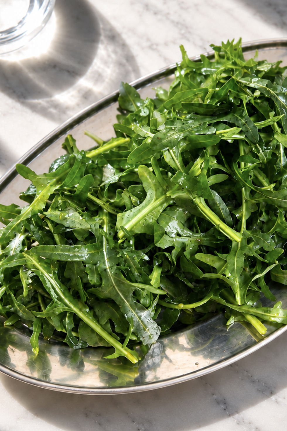 Fresh arugula leaves piled in a metal bowl on a sunlit marble counter, glistening with water droplets. Rocket Salad (Roka Salata)