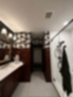 A bathroom with a deep dark red picture frame molding a little more than halfway up the wall and a b;ack and white bold wallpaper with a flying crane print on the upper part of the wall. A large walk-in shower with white tile is show on the right. A long vanity with wooden scalloped morrior is shown to the left. There is a matte black hexagon tile accent wall behind the vanity mirrors. The floor is a black and white hexagon penny tile.