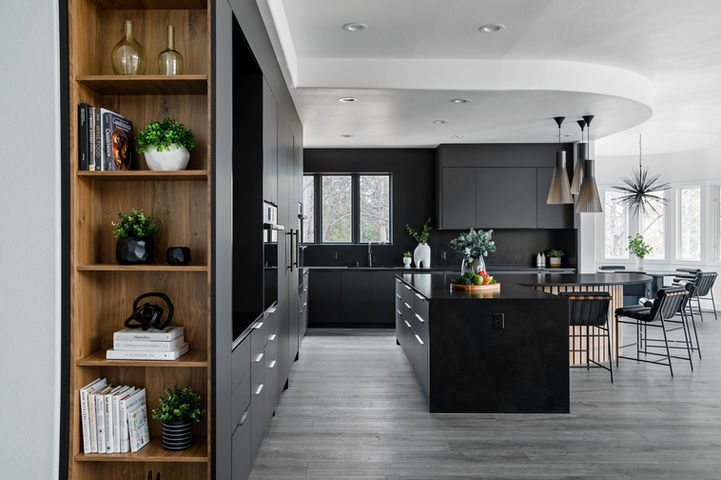 Open-concept kitchen with matte black cabinets, black stone island, sculptural pendant lights, curved white ceiling detail, walnut bookcase with plants and books, black bar stools, and bright dining nook with spiky chandelier.