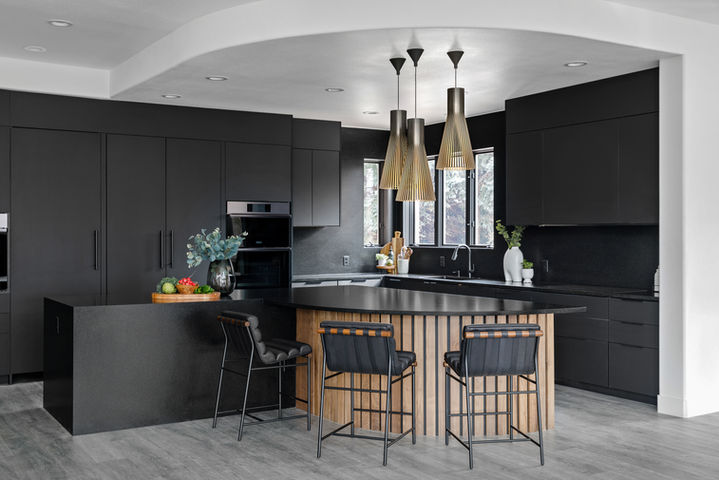 Modern black kitchen with matte cabinetry, double ovens, a textured black island with slatted wood detail, black leather bar stools, sculptural pendant lights, and a window letting in natural light.