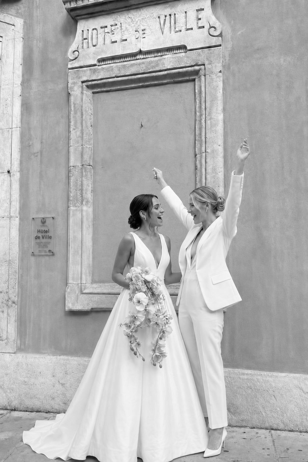 Two brides celebrating outside a town hall, one wearing a custom deep-V satin A-line wedding dress and the other wearing a modern white bridal suit by Studio RÉN.