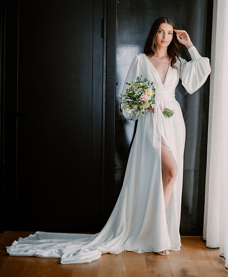 Bride standing indoors in a white robe with a thigh-high slit, holding a bouquet, with a long white train pooling on the wood