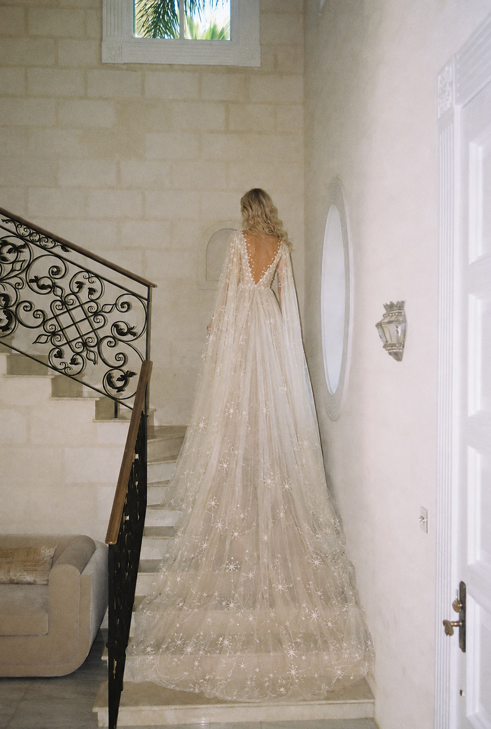 A blonde bride ascends a stone staircase in an open-back, warm-ivory tulle wedding dress with flowing cape sleeves and a cathedral-length train embroidered with stars and constellation lines. No separate veil is worn. The extended embroidered tulle creates the veil-like effect.