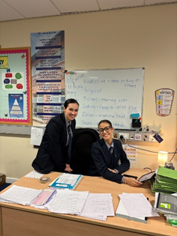 Two school girls at the teachers desk in a classroom, one sat at the computer and the other stood behind, both smiling at the camera.