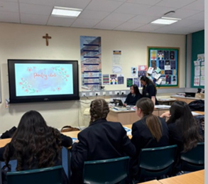 Students seated in rows looking towards interactive board that reads 'Poetry Club.' To thr right of this is two school girls at a desk presenting.