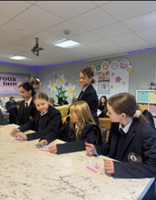Four young high school students sat at a desk facing the same direction, writing on pieces of paper. Behind them is an older student who is overseeing their work.