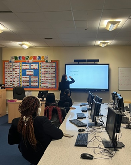 Students sat at desks with computers that are all looking towards the top of the classroom where an older student is writing on the interactive whiteboard.