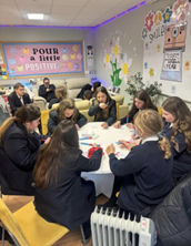 A large group of students say round a circular table writing on pieces of paper. I the background there are more students on sofas against that wall.