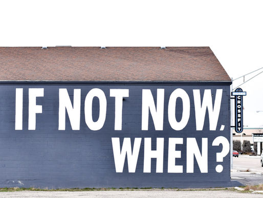 Building with blue wall displays bold white text "IF NOT NOW, WHEN?" in urban setting, with a sign on the side.