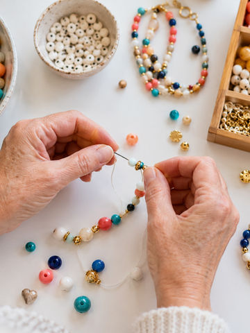 Hands stringing a beaded bracelet beside trays of beads