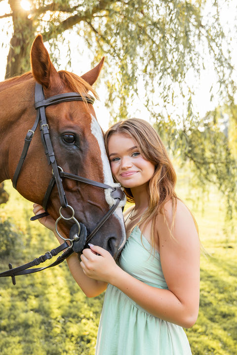 Senior girls smiling and hugging her horse under willow tree during golden hour during her senior session in Oakland Twp Mi