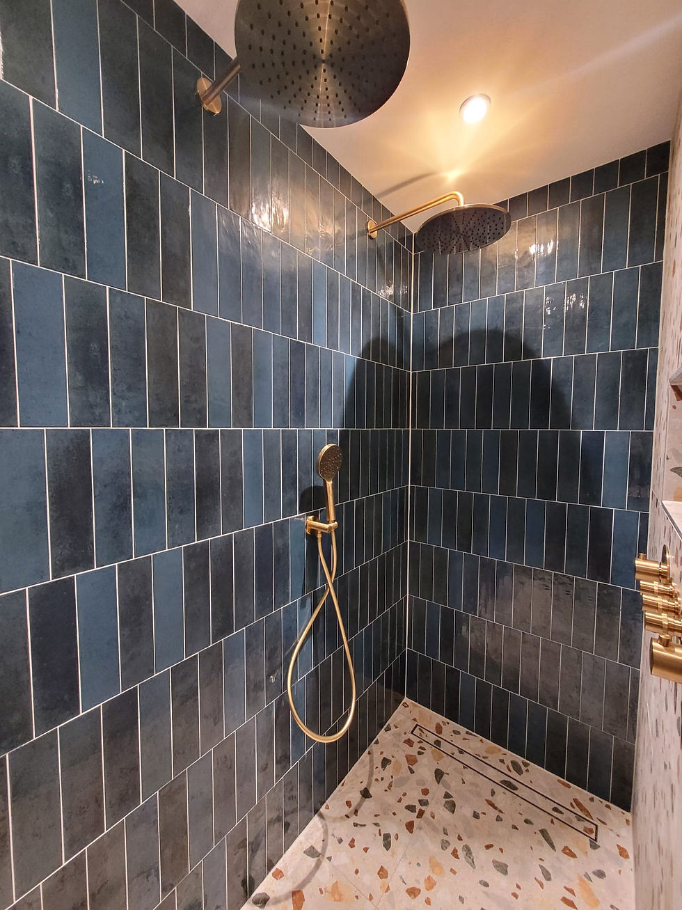 Inloopdouche met blauwe verticale wandtegels, terrazzo douchevloer en geborsteld gouden regendoucheset.
