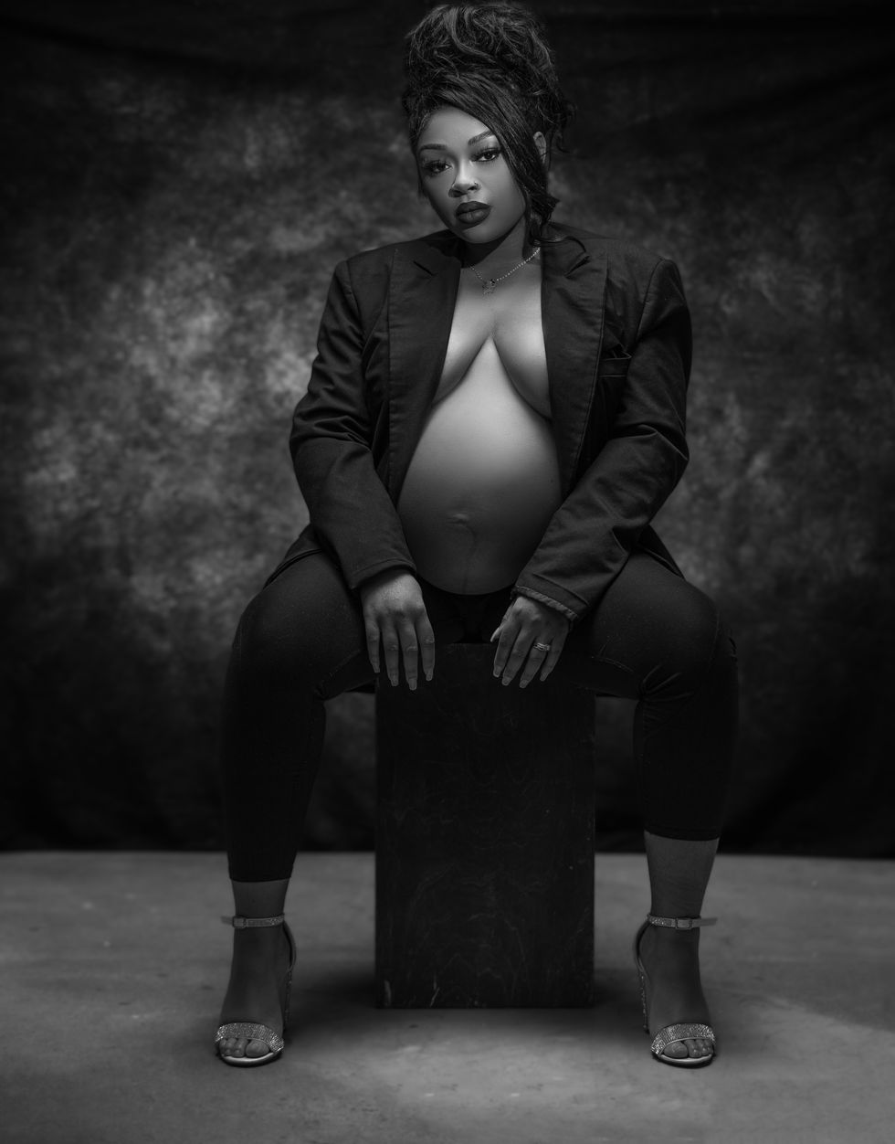 Black-and-white image of a pregnant Black woman in an open black blazer and heels, seated and staring directly at the camera — bold maternity portrait by Shooters Society Studios.