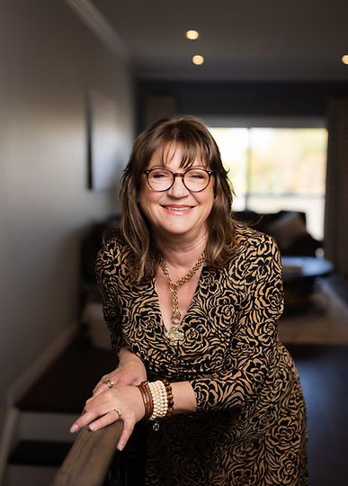 Linda Bischoff, Avalon Lane Studio, Workplace Design Consultant and Wellness Guide. Linda stands in her hallway, wearing a tan printed dress with her hands on a railing.