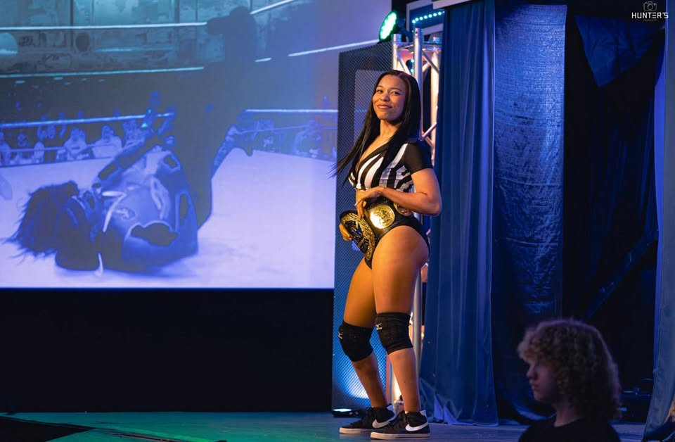 Confident female wrestler posing on stage with championship belts, blue lights, and a wrestling match playing on a large screen પાછળ