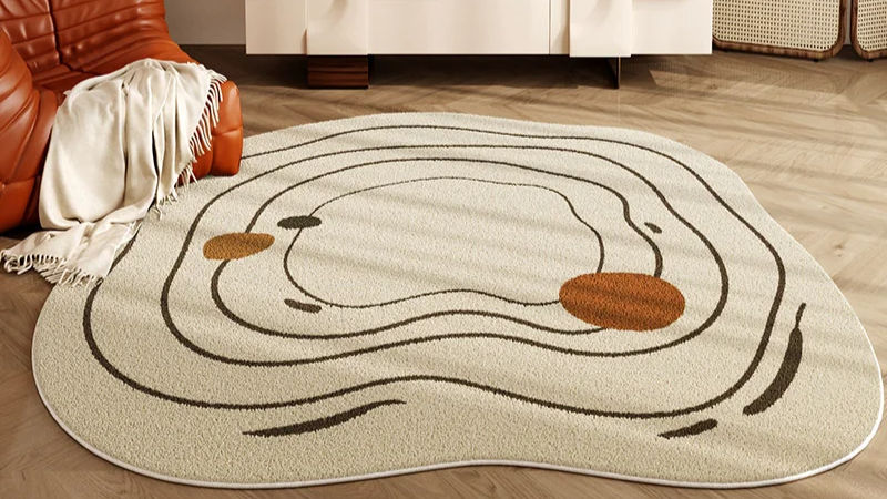 A beige rug with abstract black and brown swirl patterns on a wooden floor. A brown chair with a white blanket sits in the corner.