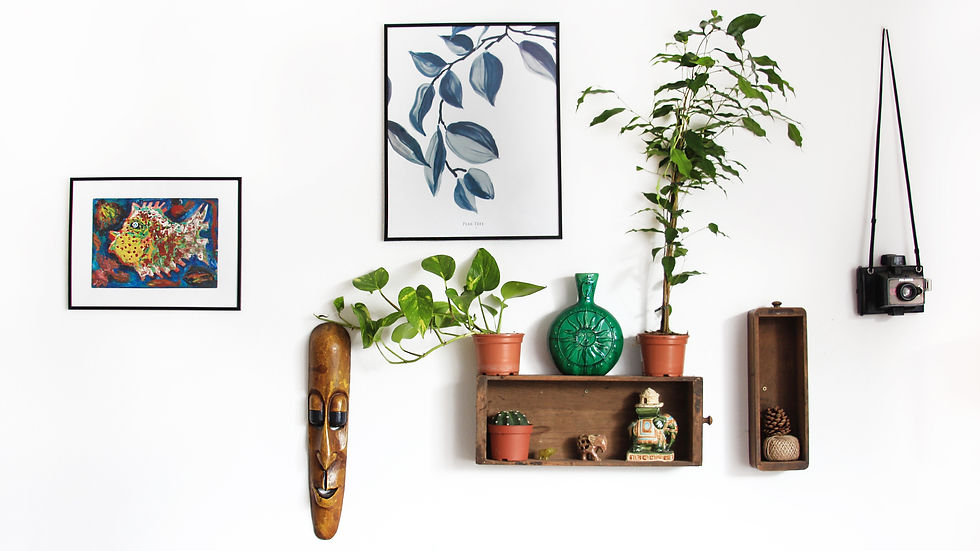 Wall with art and plants: colorful fish painting, leaf print, wooden mask, green vase, potted plants, vintage camera, and small sculptures.