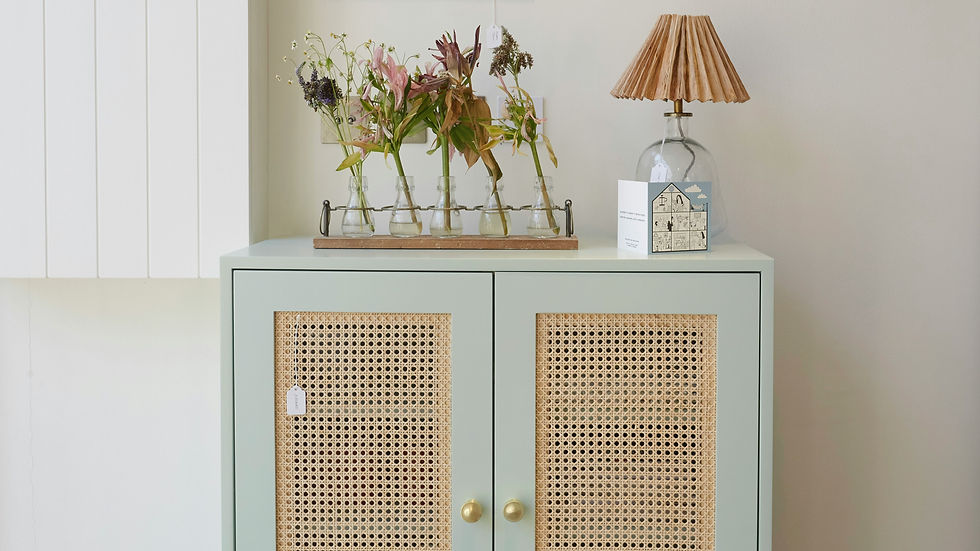 Light green cabinet with rattan doors, topped with an arrangement of flowers in glass vases, a card, and a lamp with a pleated shade.