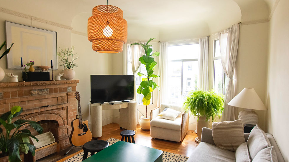 Cozy living room with a beige sofa, guitar by a brick fireplace, large plants, TV, and woven light fixture. Bright, airy, and inviting.