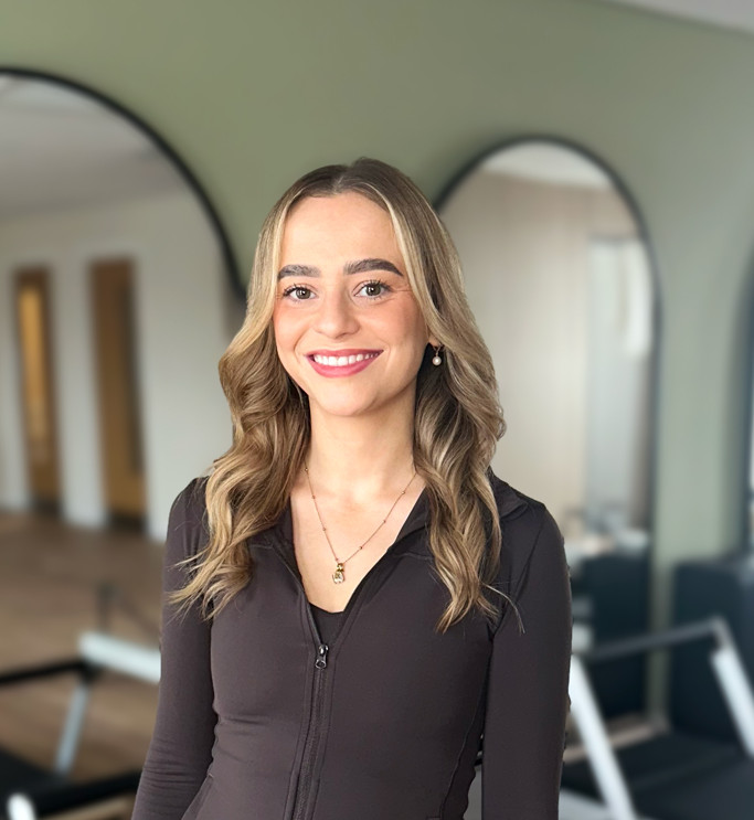 Professional headshot of Kate, Renu Studios instructor and owner standing in front of reformer machines in the Studio