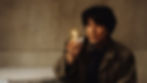 Takabe from CUre (1977) in a trench coat holds a lit lighter, focused expression. Dimly lit room with a beige wall; mood is tense and contemplative.
