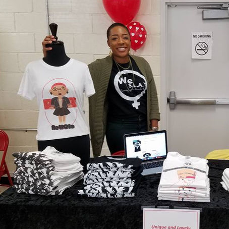 A woman smiles behind a table with folded T-shirts and a laptop. A mannequin displays a design. Red heart balloon in background. Sign reads "Unique and Lovely".
