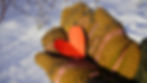 Hands in brown mittens hold a red paper heart in a snowy setting. Warm, cozy vibe with blurred snow-covered background.
