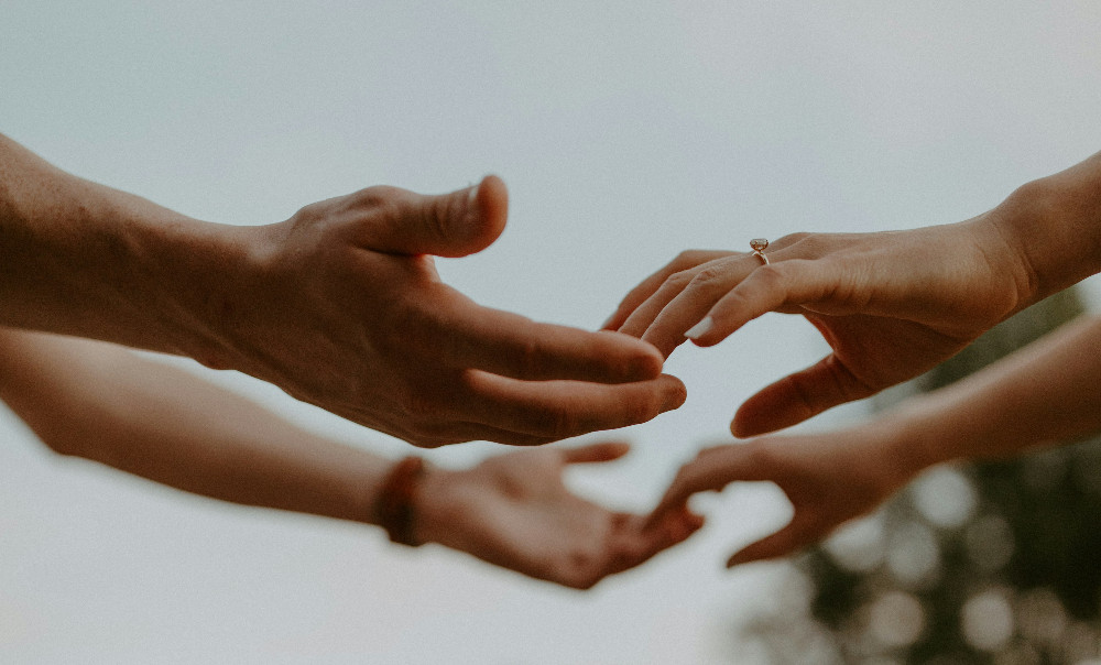 Four hands reach toward each other outdoors against a blurred sky, one wearing a ring, suggesting love and connection.