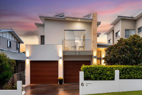 Modern, two-story home with contemporary design, featuring a balcony and double garage. Surrounded by greenery and a low brick wall.