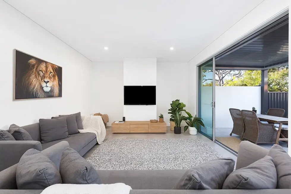 Modern living room with gray sectional sofa, rug, and wall-mounted TV. Sliding glass door leads to patio with dining area. Lion print hangs on the wall.
